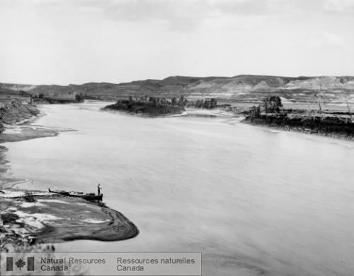 Photo 1090 : T.C. Weston's boats on the Red Deer River, Alberta. Photo looks down on river and surrounding landscape. On the right a man is fishing from the  ...