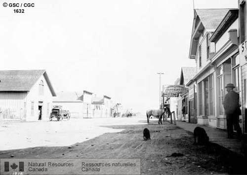 Photo 1632 : MacLeod (Alberta). Vu de la rue principale en terre, les trottoirs de bois et les devantures de magasins en bois. Il y a des chariots, des chevaux et  ...
