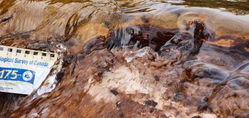 Photo 2022-458 : Field photo of the Fe-oxyhydroxide that has precipitated on the stream bed. Note the rhythmic layering of lighter and darker brown layers of  ...