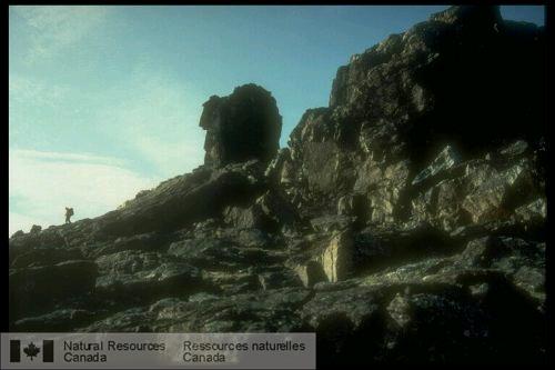 Photo EMR-11104 : Silouette intéressante d'un affleurement et d'un géologue revenant d'un levé ( gauche de l'affleurement). Vue vers le sud-ouest, au nord de la baie  ...